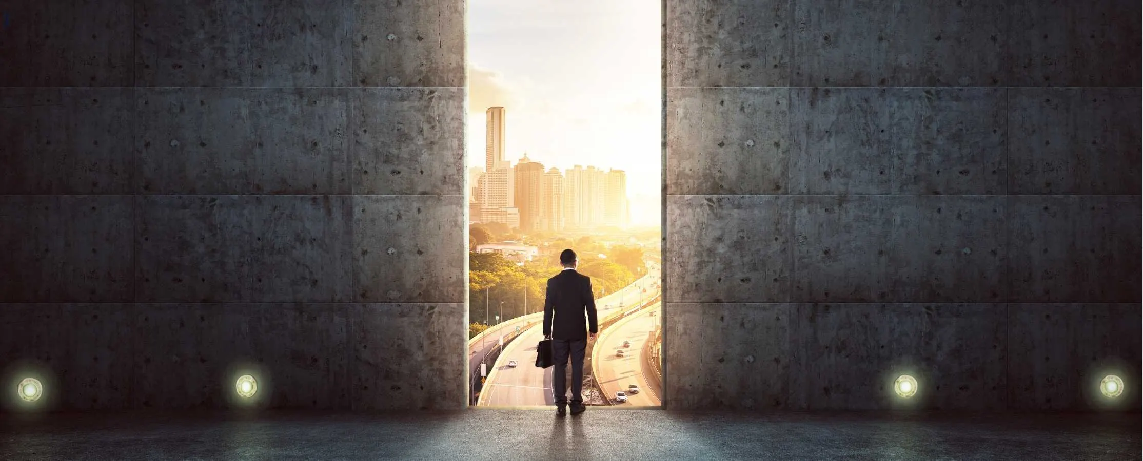 Man with a briefcase walking through a gap in a concrete wall towards a sunlit city