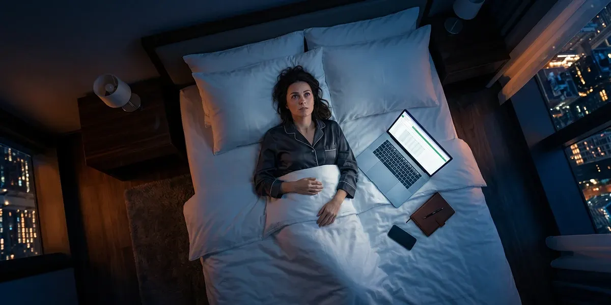 Frau liegt wach im Bett, neben ihr ein aufgeklappter Laptop und ihr Handy